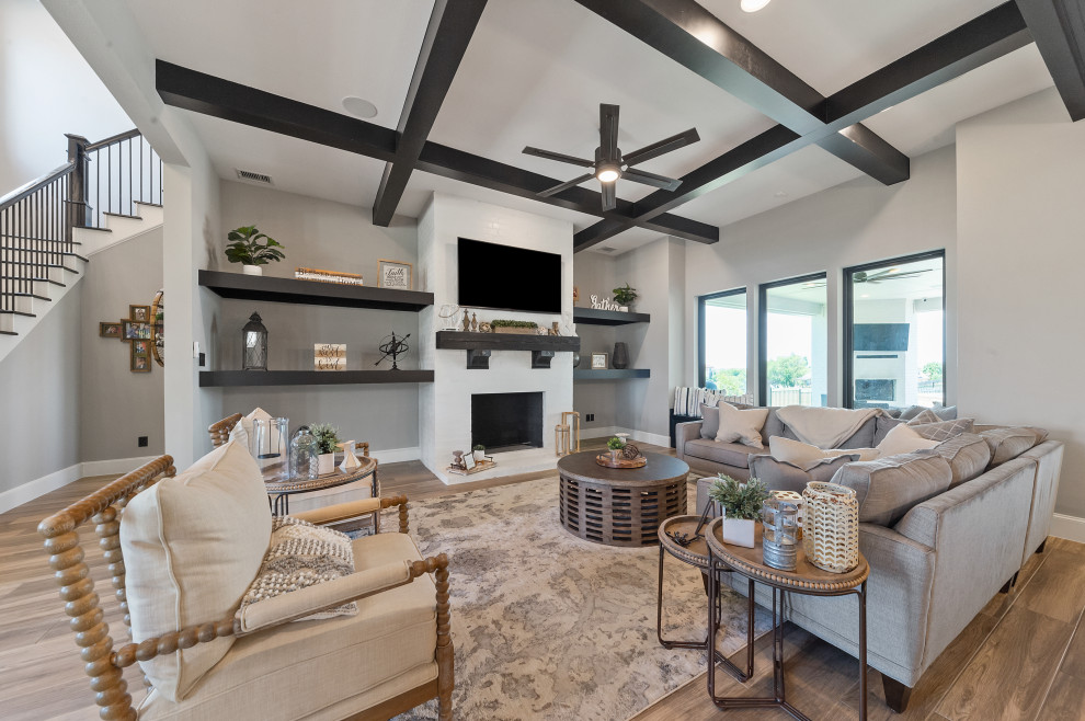 This is an example of a large farmhouse open plan games room in Dallas with grey walls, medium hardwood flooring, a wood burning stove, a brick fireplace surround, a wall mounted tv, brown floors and exposed beams.