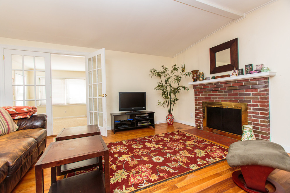 Modelo de sala de estar abierta clásica de tamaño medio con paredes beige, suelo de madera en tonos medios, todas las chimeneas, marco de chimenea de ladrillo y televisor independiente