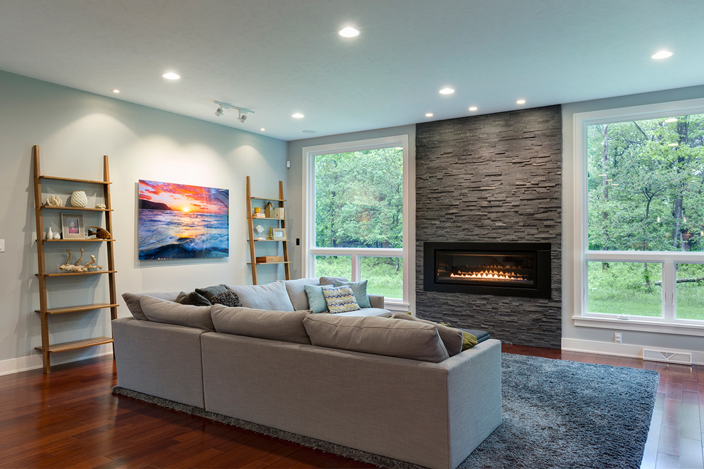 Modelo de sala de estar abierta contemporánea grande con paredes grises, suelo de madera en tonos medios, todas las chimeneas, marco de chimenea de piedra y televisor colgado en la pared