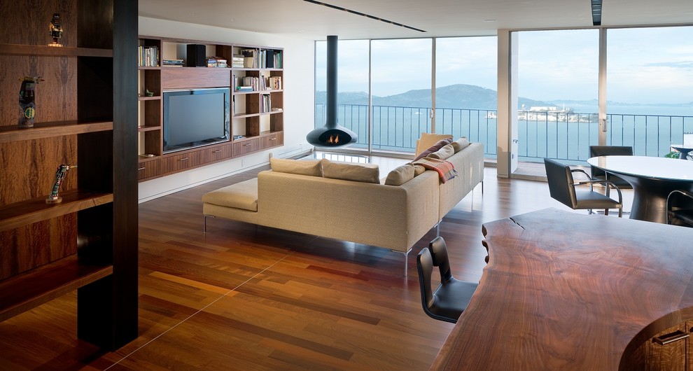 Réalisation d'un salon design ouvert avec un mur blanc, un sol en bois brun, cheminée suspendue et un téléviseur encastré.