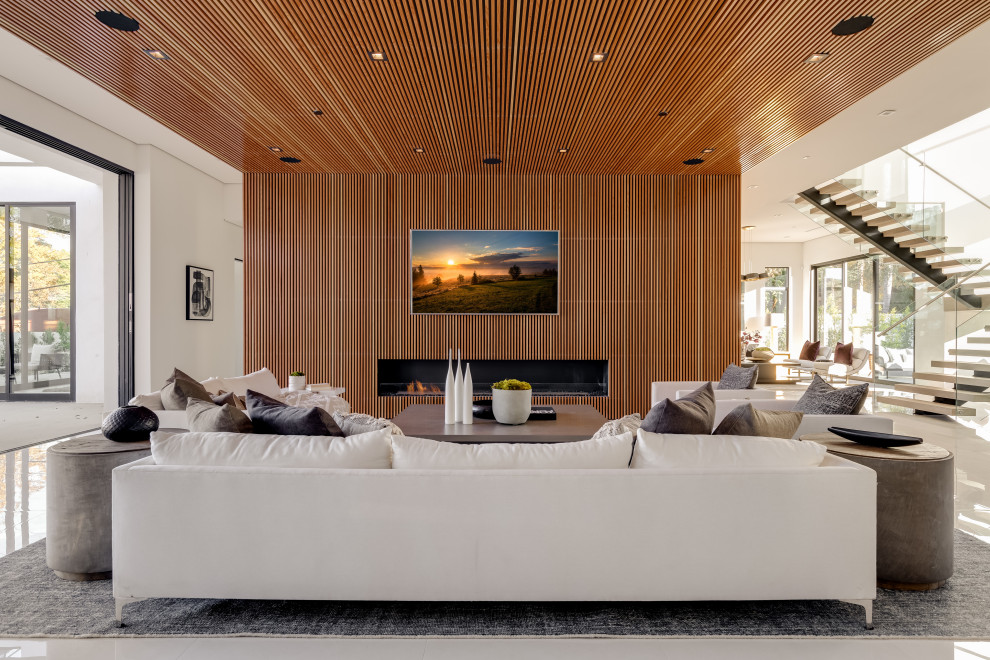 Ejemplo de sala de estar abierta contemporánea extra grande con paredes marrones, suelo de baldosas de porcelana, todas las chimeneas, marco de chimenea de madera, televisor colgado en la pared y suelo blanco