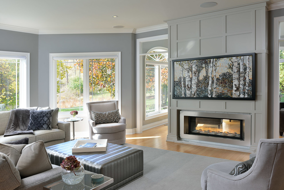 Modelo de sala de estar clásica renovada grande con paredes grises, suelo de madera clara, chimenea de doble cara, marco de chimenea de madera y televisor colgado en la pared
