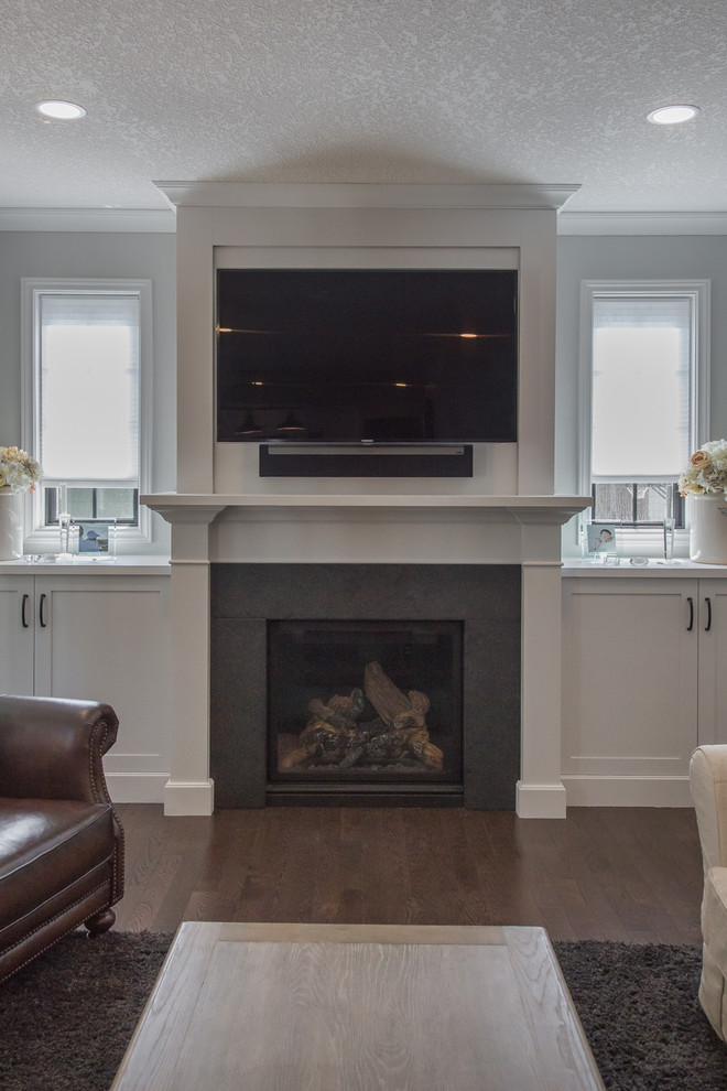 This is an example of a medium sized traditional open plan games room in Toronto with white walls, dark hardwood flooring, a standard fireplace, a wooden fireplace surround, a wall mounted tv and brown floors.