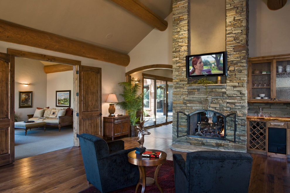 Inspiration for a large rustic enclosed games room in Other with a home bar, beige walls, medium hardwood flooring, a two-sided fireplace, a stone fireplace surround and a wall mounted tv.