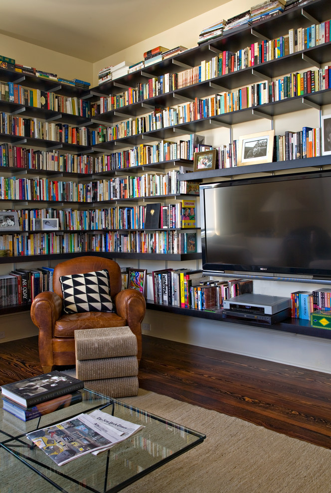 Imagen de sala de estar con biblioteca actual con paredes beige, suelo de madera oscura, televisor colgado en la pared y alfombra