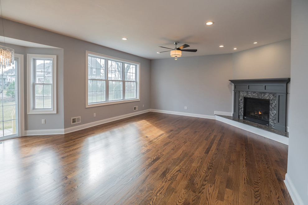 Modelo de sala de estar clásica renovada con paredes grises, suelo de madera clara, chimenea de esquina, marco de chimenea de piedra y suelo marrón