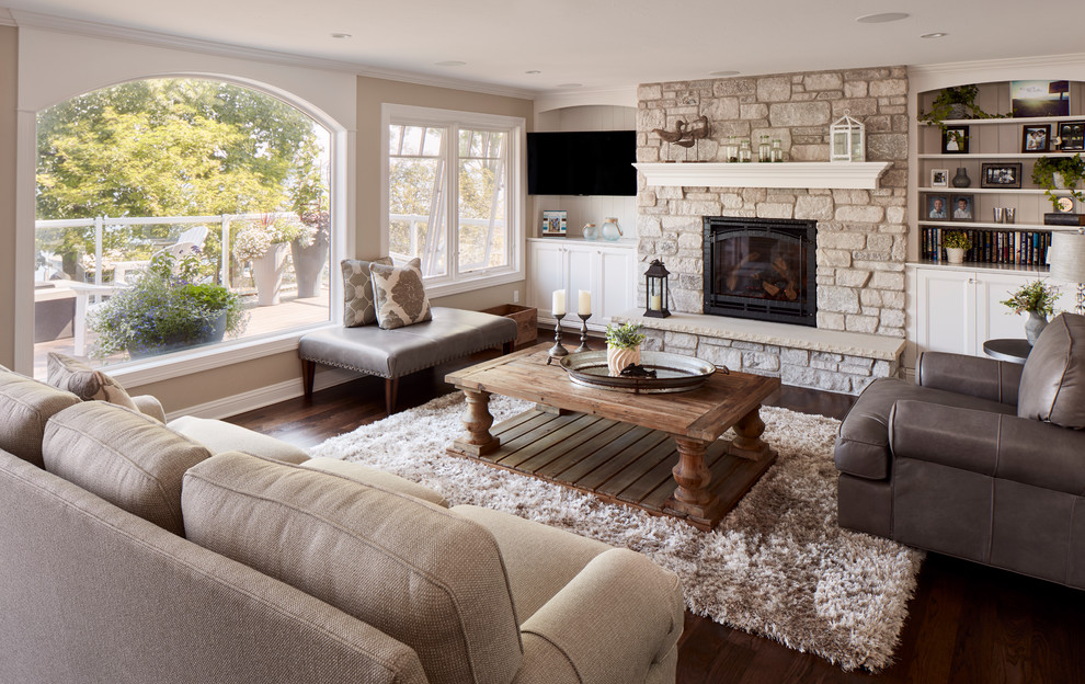 Modelo de sala de estar costera con paredes beige, suelo de madera oscura, todas las chimeneas, marco de chimenea de piedra, televisor colgado en la pared y alfombra