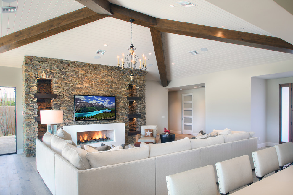 Imagen de sala de estar abierta de estilo americano grande con paredes blancas, suelo de madera clara, marco de chimenea de piedra, televisor colgado en la pared, suelo marrón y chimenea lineal