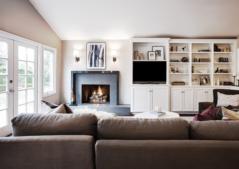Cette image montre une grande salle de séjour design ouverte avec une cheminée standard, un téléviseur encastré, un manteau de cheminée en pierre, un mur gris et un sol marron.
