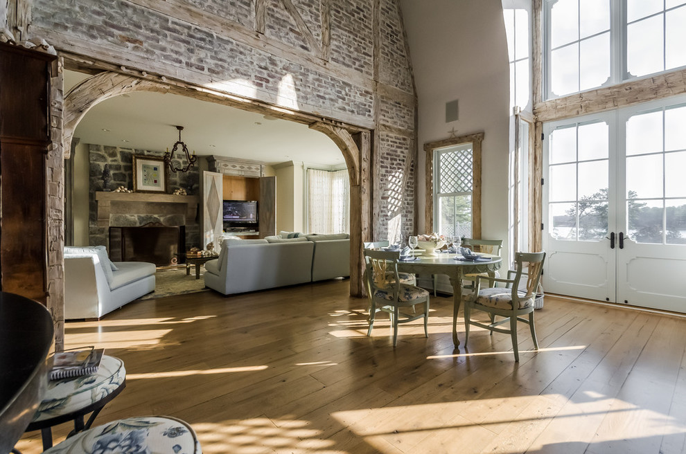 Modelo de sala de estar abierta campestre grande con paredes blancas, suelo de madera en tonos medios, todas las chimeneas y marco de chimenea de piedra