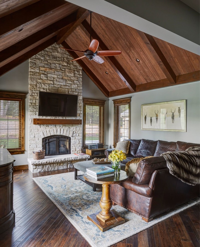 Ejemplo de sala de estar de estilo americano de tamaño medio con paredes grises, suelo de madera oscura, todas las chimeneas, marco de chimenea de piedra y televisor colgado en la pared