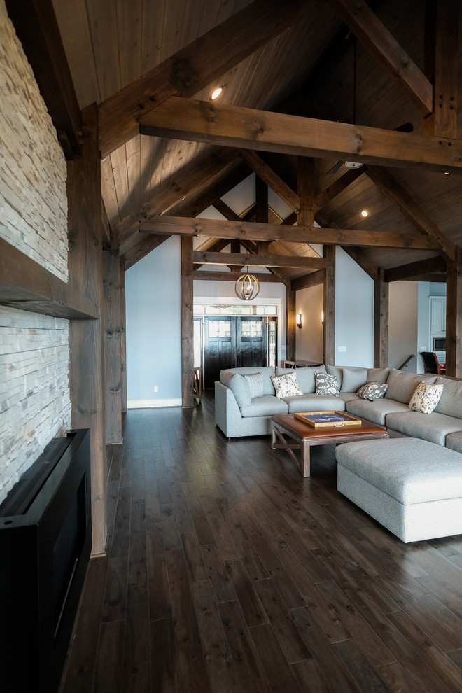 Modelo de sala de estar abierta rural grande con paredes grises, suelo de madera oscura, chimenea lineal, marco de chimenea de piedra y suelo marrón
