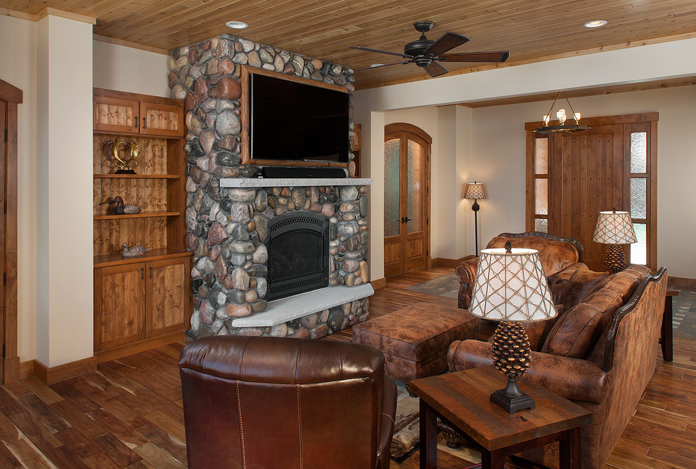 This is an example of a medium sized rustic open plan games room in Grand Rapids with beige walls, a standard fireplace, a stone fireplace surround, a wall mounted tv and medium hardwood flooring.