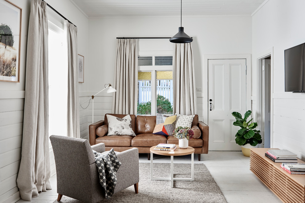 На фото: гостиная комната в скандинавском стиле с белыми стенами, деревянным полом, телевизором на стене, белым полом и коричневым диваном без камина с