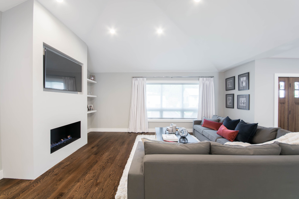 Modelo de sala de estar abierta moderna grande con paredes grises, suelo de madera oscura, chimenea lineal, marco de chimenea de yeso, televisor colgado en la pared y suelo marrón