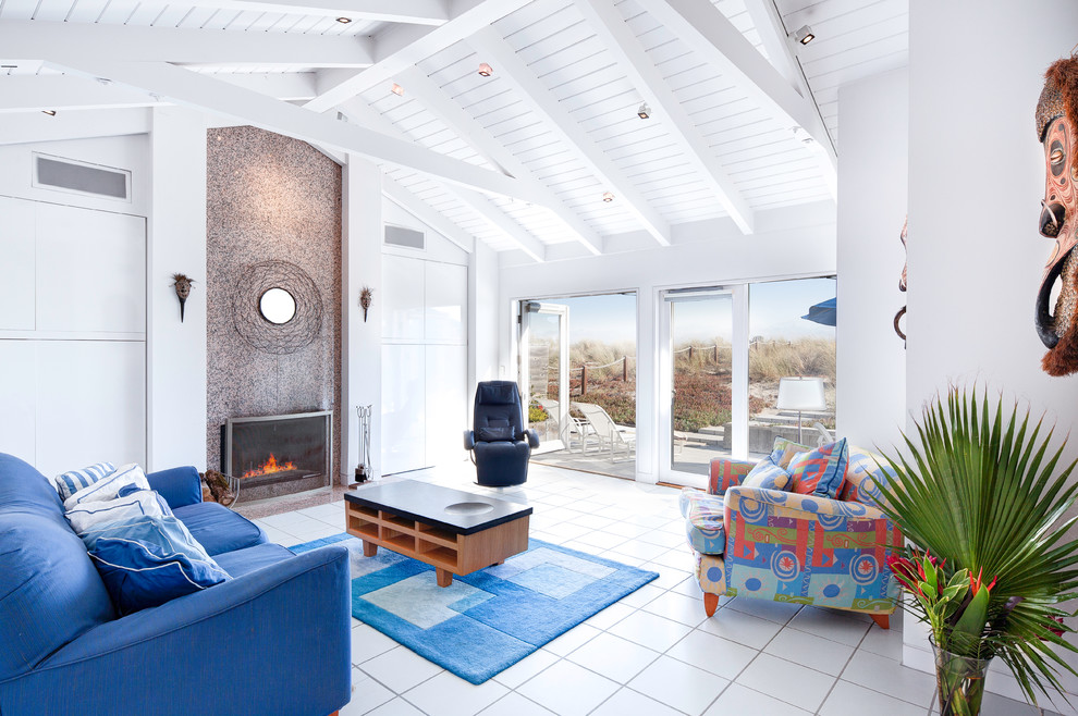 На фото: большая открытая гостиная комната в морском стиле с белыми стенами, полом из керамогранита, стандартным камином, фасадом камина из камня, белым полом и синим диваном