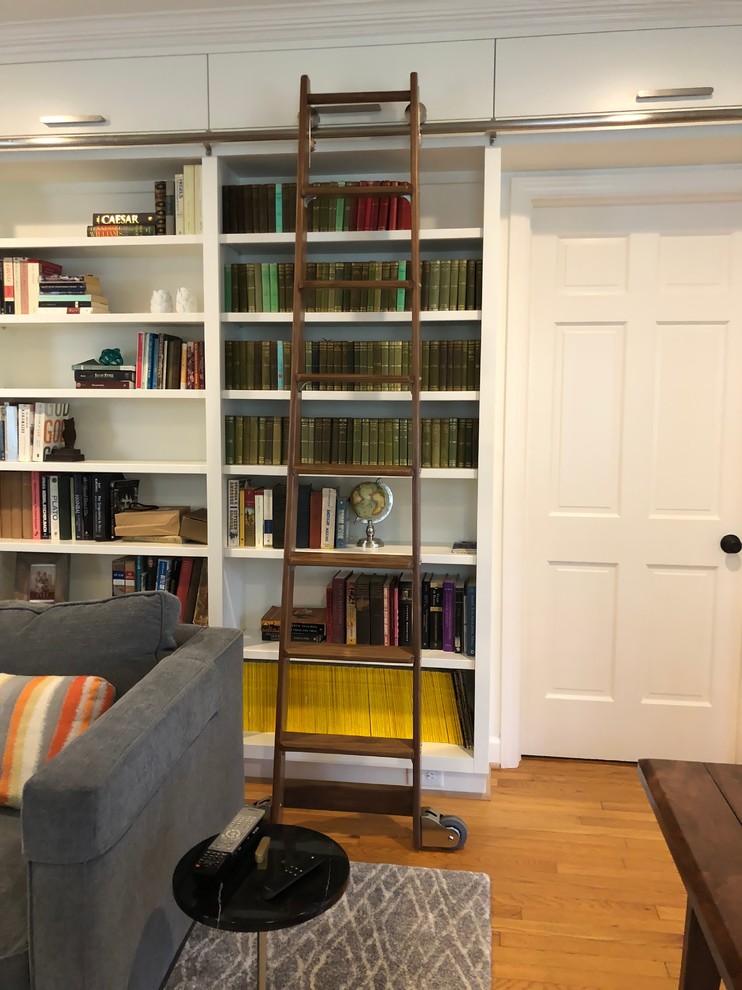 На фото: большая изолированная гостиная комната в стиле неоклассика (современная классика) с с книжными шкафами и полками, белыми стенами, светлым паркетным полом, скрытым телевизором и коричневым полом без камина