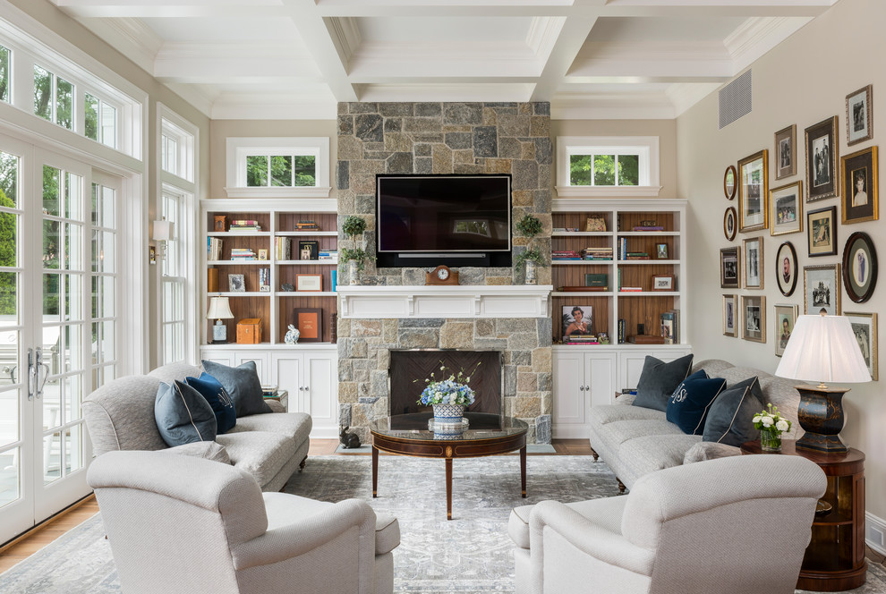 На фото: гостиная комната в классическом стиле с с книжными шкафами и полками, бежевыми стенами, стандартным камином, фасадом камина из камня, телевизором на стене и ковром на полу