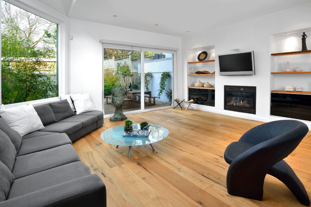 Modelo de sala de estar abierta contemporánea con paredes blancas, todas las chimeneas, televisor colgado en la pared, suelo de madera clara y suelo beige