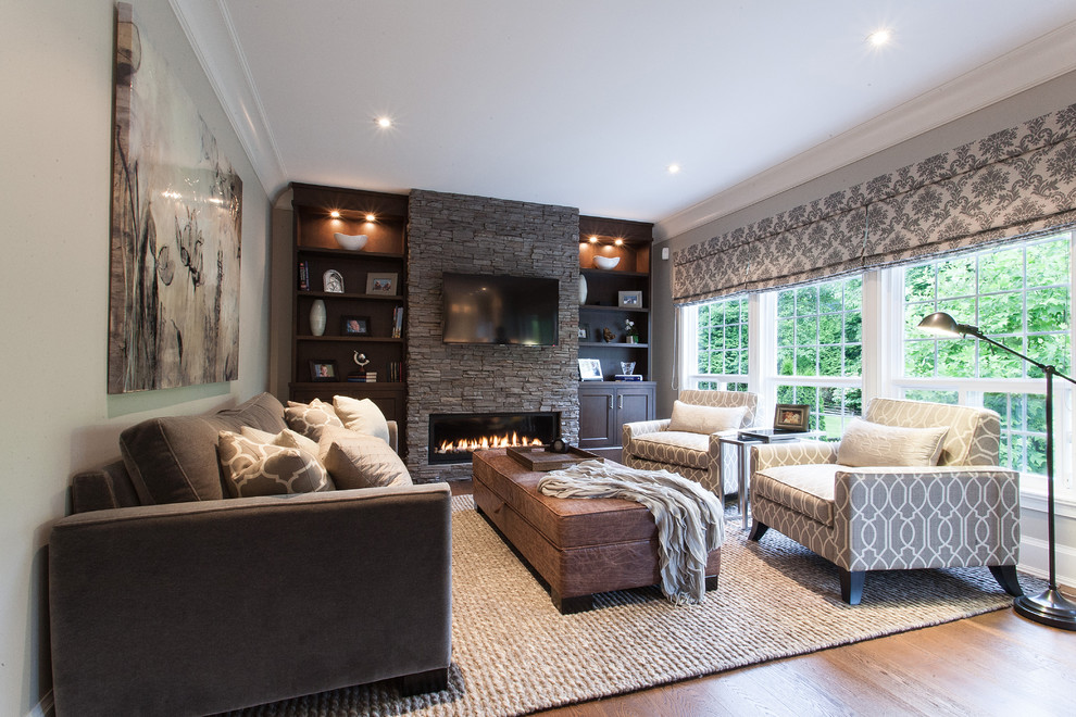 Modelo de sala de estar clásica grande con paredes grises, suelo de madera en tonos medios, chimenea lineal, marco de chimenea de piedra y televisor colgado en la pared