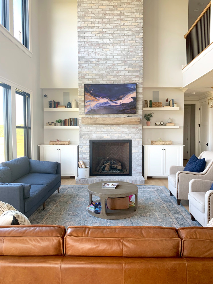 Modelo de sala de estar abierta de estilo de casa de campo grande con paredes grises, suelo de madera clara, todas las chimeneas, marco de chimenea de ladrillo y televisor colgado en la pared