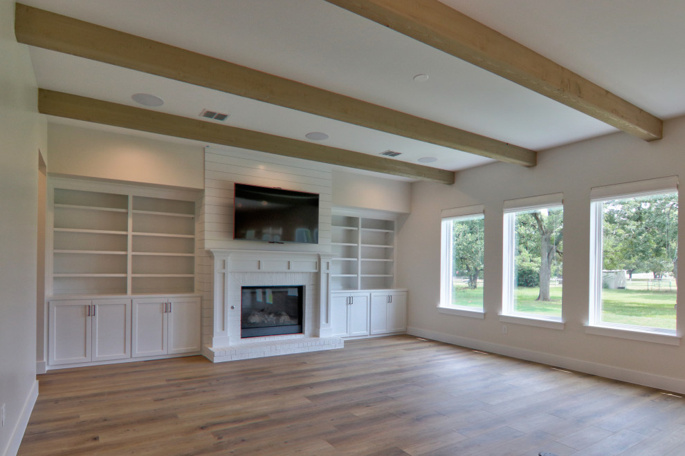 На фото: большая открытая гостиная комната в стиле кантри с серыми стенами, полом из винила, стандартным камином, фасадом камина из дерева, телевизором на стене и коричневым полом