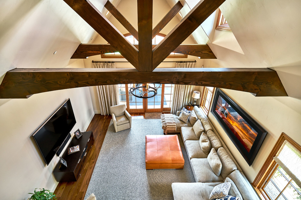 Modelo de sala de estar abierta campestre extra grande con paredes beige, suelo de madera oscura y televisor colgado en la pared
