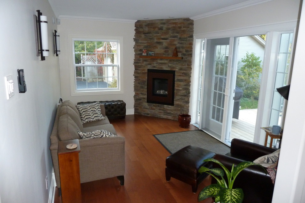 Modelo de sala de estar tipo loft contemporánea pequeña con paredes grises, suelo de madera en tonos medios, chimenea de esquina, marco de chimenea de piedra y pared multimedia
