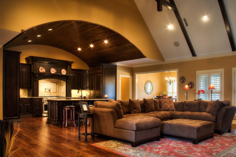 Modelo de sala de estar abierta de estilo americano grande con televisor colgado en la pared, paredes marrones, suelo de madera en tonos medios, todas las chimeneas y marco de chimenea de piedra