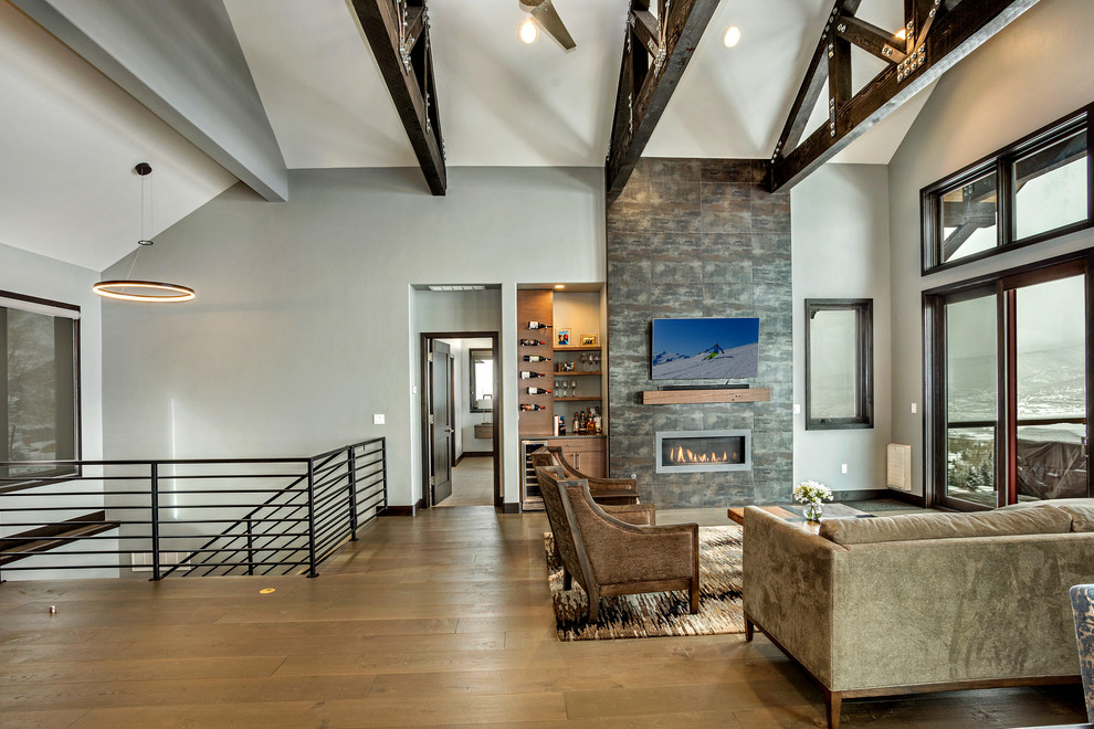 Inspiration for a medium sized modern open plan games room in Denver with a home bar, grey walls, medium hardwood flooring, a standard fireplace, a stone fireplace surround, a wall mounted tv and brown floors.