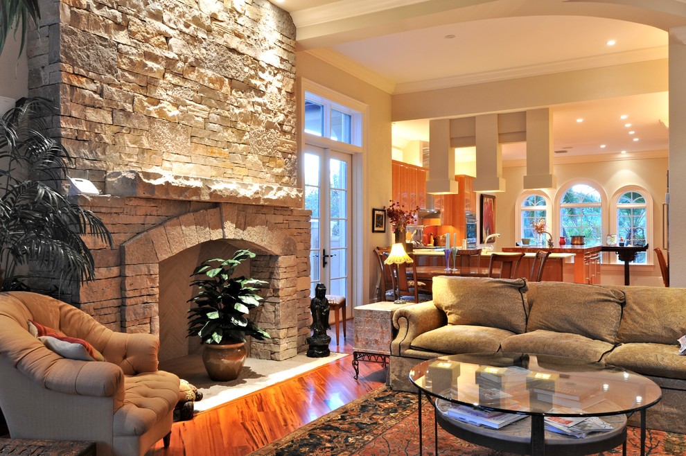 Cette image montre une grande salle de séjour traditionnelle ouverte avec un mur beige, un sol en bois brun, une cheminée standard et un manteau de cheminée en pierre.