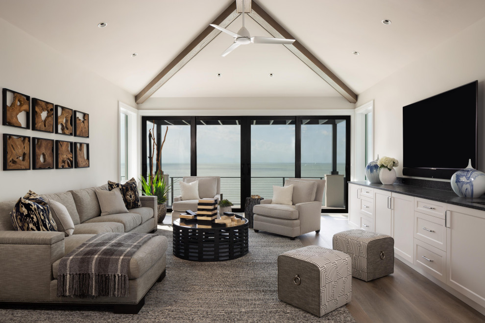 Imagen de sala de estar abovedada costera grande sin chimenea con televisor colgado en la pared, paredes blancas, suelo de madera oscura y vigas vistas