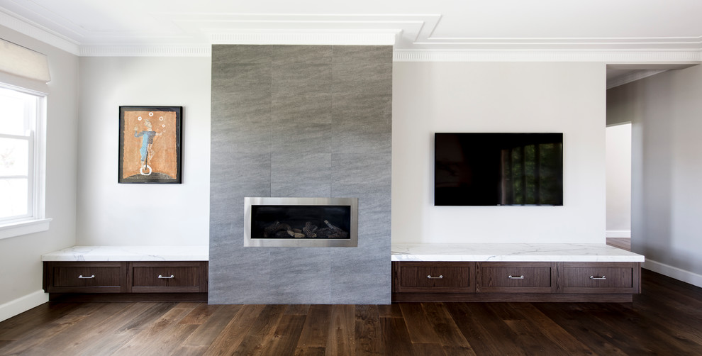На фото: большая открытая гостиная комната в стиле неоклассика (современная классика) с серыми стенами, паркетным полом среднего тона, стандартным камином, фасадом камина из плитки, телевизором на стене и коричневым полом