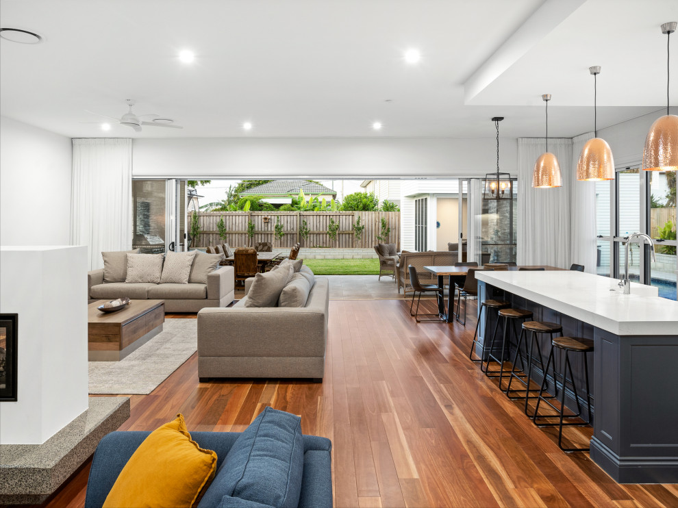 This is an example of a medium sized coastal open plan games room in Brisbane with white walls, medium hardwood flooring, a two-sided fireplace, a plastered fireplace surround, a wall mounted tv and multi-coloured floors.