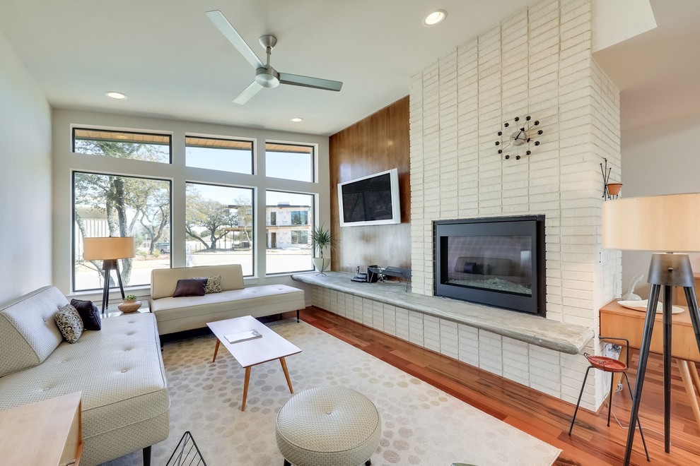 На фото: открытая гостиная комната в стиле ретро с белыми стенами, паркетным полом среднего тона, фасадом камина из кирпича, телевизором на стене, стандартным камином, коричневым полом и ковром на полу