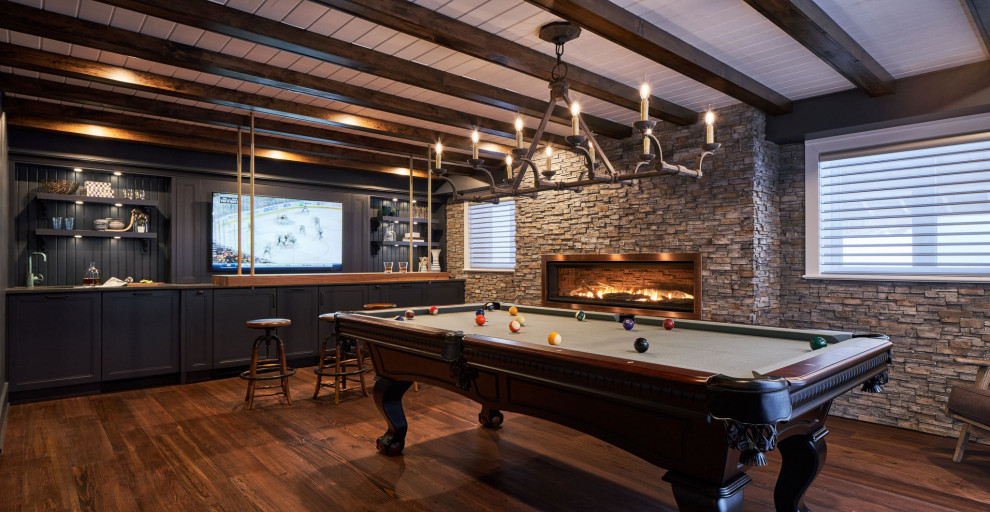This is an example of a rustic games room in Ottawa with a game room, grey walls, dark hardwood flooring, a standard fireplace, a stone fireplace surround, a wall mounted tv, brown floors and exposed beams.