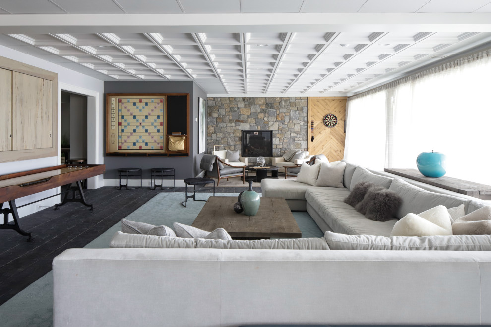 Пример оригинального дизайна: открытая комната для игр среднего размера в стиле неоклассика (современная классика) с серыми стенами, деревянным полом, стандартным камином, фасадом камина из камня, мультимедийным центром и черным полом