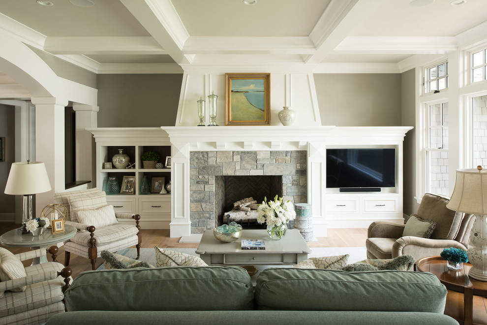 Modelo de sala de estar costera con paredes grises, suelo de madera en tonos medios, todas las chimeneas, marco de chimenea de piedra, televisor colgado en la pared y suelo marrón