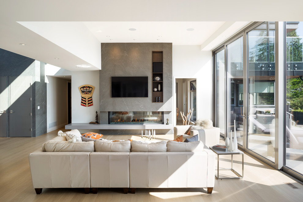 Ejemplo de sala de estar abierta actual grande con paredes blancas, suelo de madera clara, chimenea de doble cara, marco de chimenea de piedra y televisor colgado en la pared