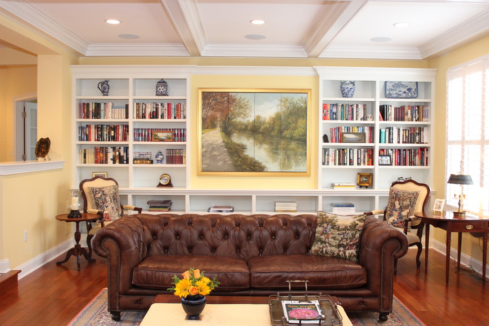 Cette photo montre une grande salle de séjour chic avec une bibliothèque ou un coin lecture, un téléviseur dissimulé, un mur jaune, un sol en bois brun et un sol marron.
