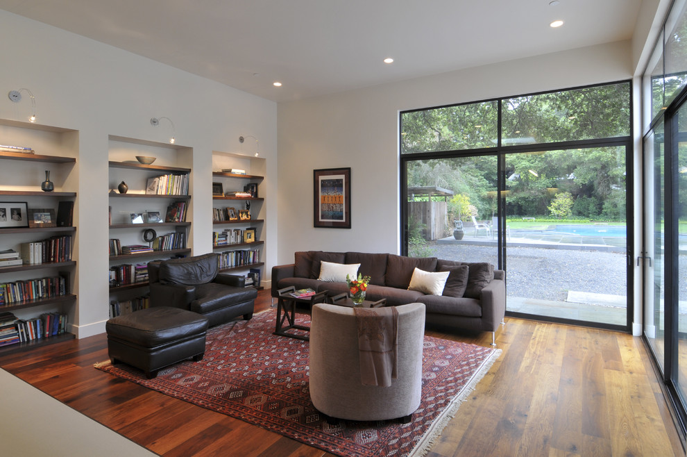 Imagen de sala de estar con biblioteca abierta actual sin chimenea con paredes blancas, suelo de madera en tonos medios y alfombra