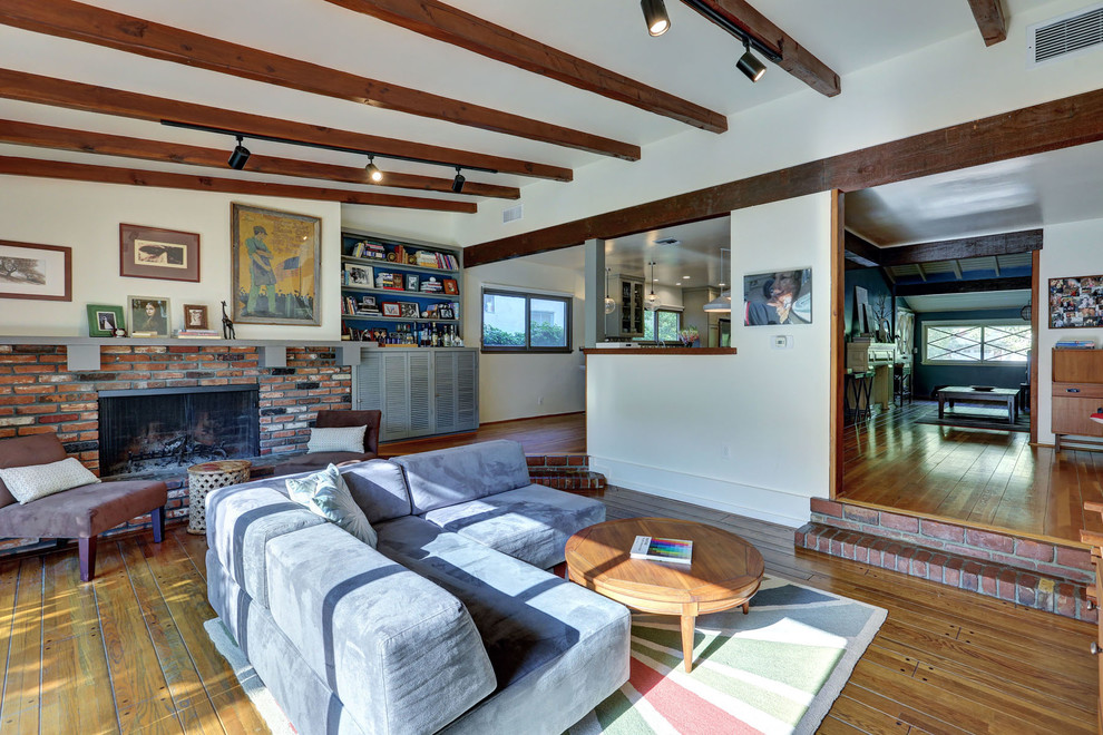 Cette image montre une salle de séjour vintage ouverte avec un sol en bois brun, une cheminée standard, un manteau de cheminée en brique et un mur blanc.