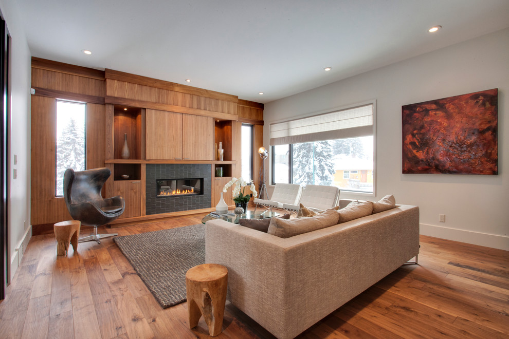 Modernes Wohnzimmer mit weißer Wandfarbe, braunem Holzboden, gefliester Kaminumrandung, verstecktem TV und Gaskamin in Calgary