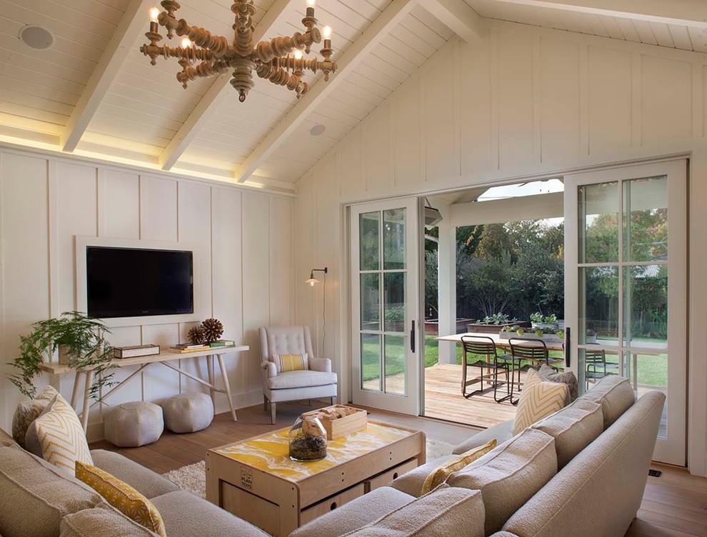 Modelo de sala de estar de estilo de casa de campo sin chimenea con paredes blancas, televisor colgado en la pared y suelo de madera en tonos medios