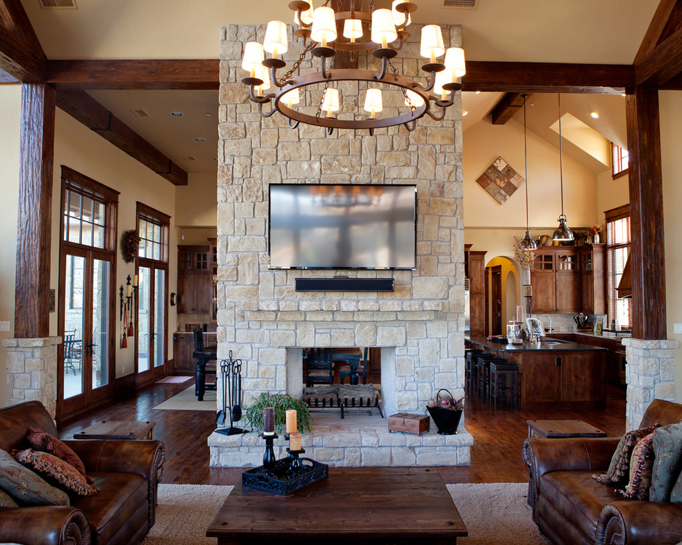 This is an example of a large rustic open plan games room in Dallas with yellow walls, dark hardwood flooring, a two-sided fireplace, a stone fireplace surround, a wall mounted tv and brown floors.