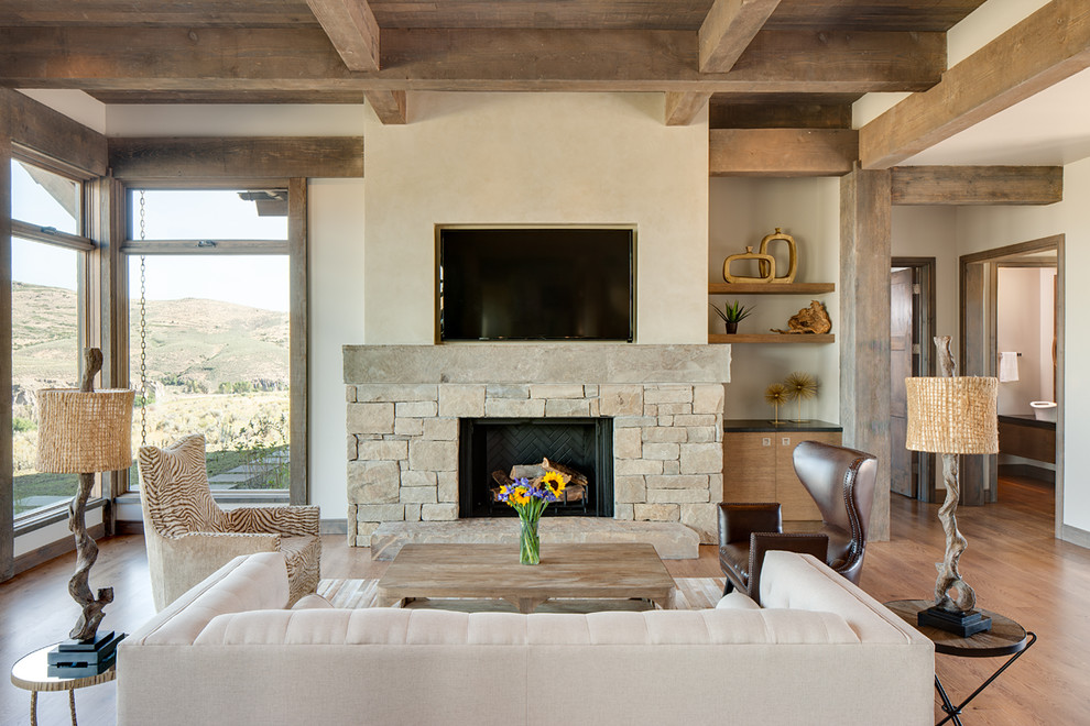 Modelo de sala de estar rural con paredes beige, suelo de madera en tonos medios, todas las chimeneas, marco de chimenea de piedra, televisor colgado en la pared y alfombra