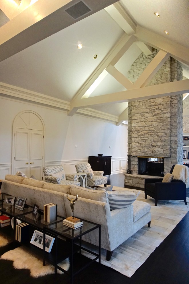 На фото: большая гостиная комната в стиле неоклассика (современная классика) с белыми стенами, темным паркетным полом, двусторонним камином и фасадом камина из камня с