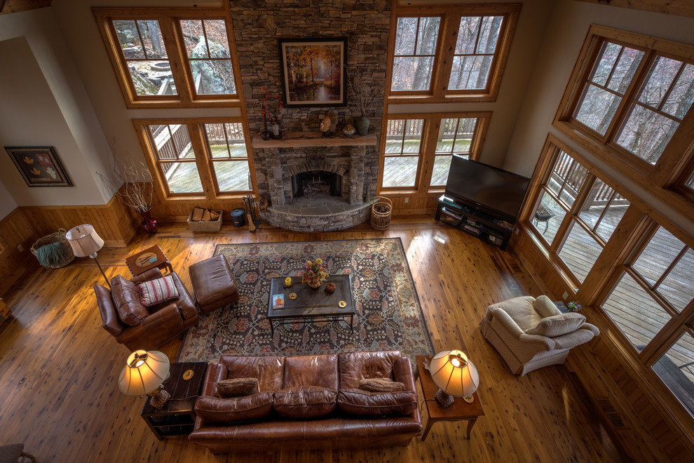 Cette image montre une grande salle de séjour chalet avec un mur beige, un sol en bois brun, une cheminée standard, un manteau de cheminée en pierre et un téléviseur indépendant.