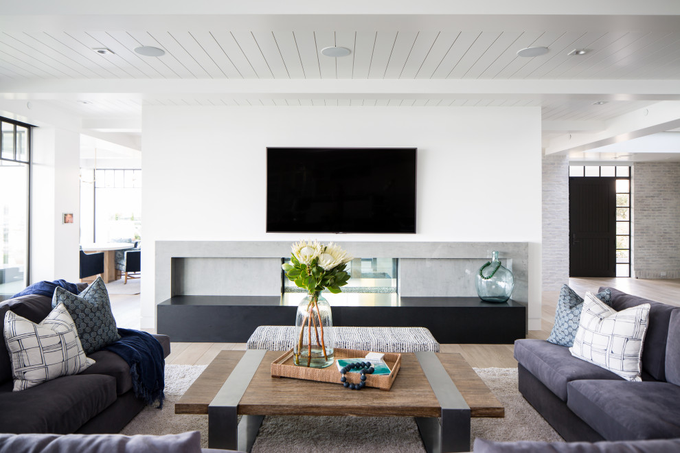 Modelo de sala de estar abierta costera extra grande con paredes blancas, suelo de madera clara, chimenea lineal y televisor colgado en la pared