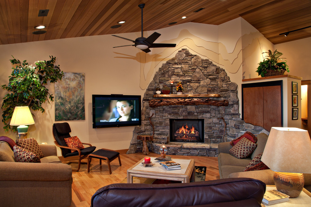 Imagen de sala de estar rural con paredes beige, suelo de madera en tonos medios, chimenea de esquina, marco de chimenea de piedra y televisor colgado en la pared